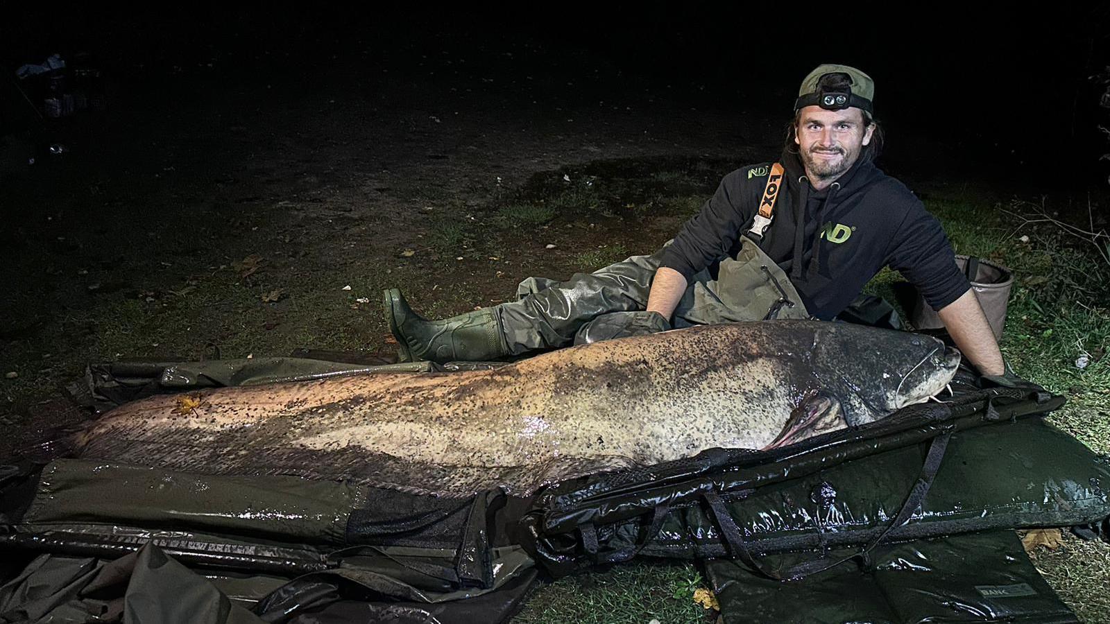 Shaun Ing, lying on the ground next to a large catfish in a night time image taken using a flash. 