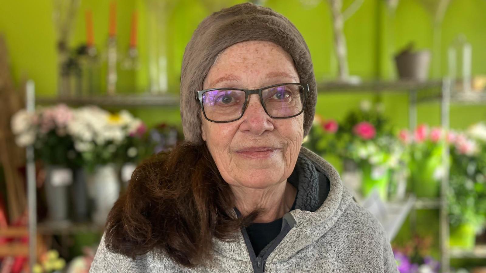 A woman wears rectangle glasses and a fluffy hat. Behind her, floral displays line a lime green wall.