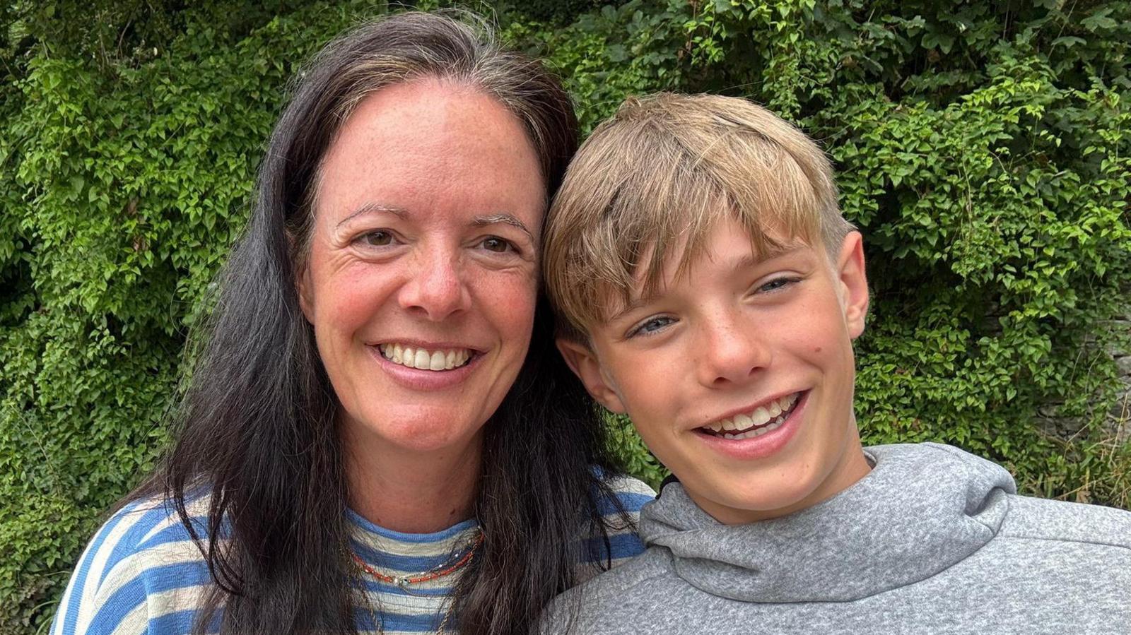 Sarah and her 11-year-old son Osian smiling at the camera. Sarah has long black hair and a striped white and blue top. Osian is wearing a grey jumper and has light brown hair.