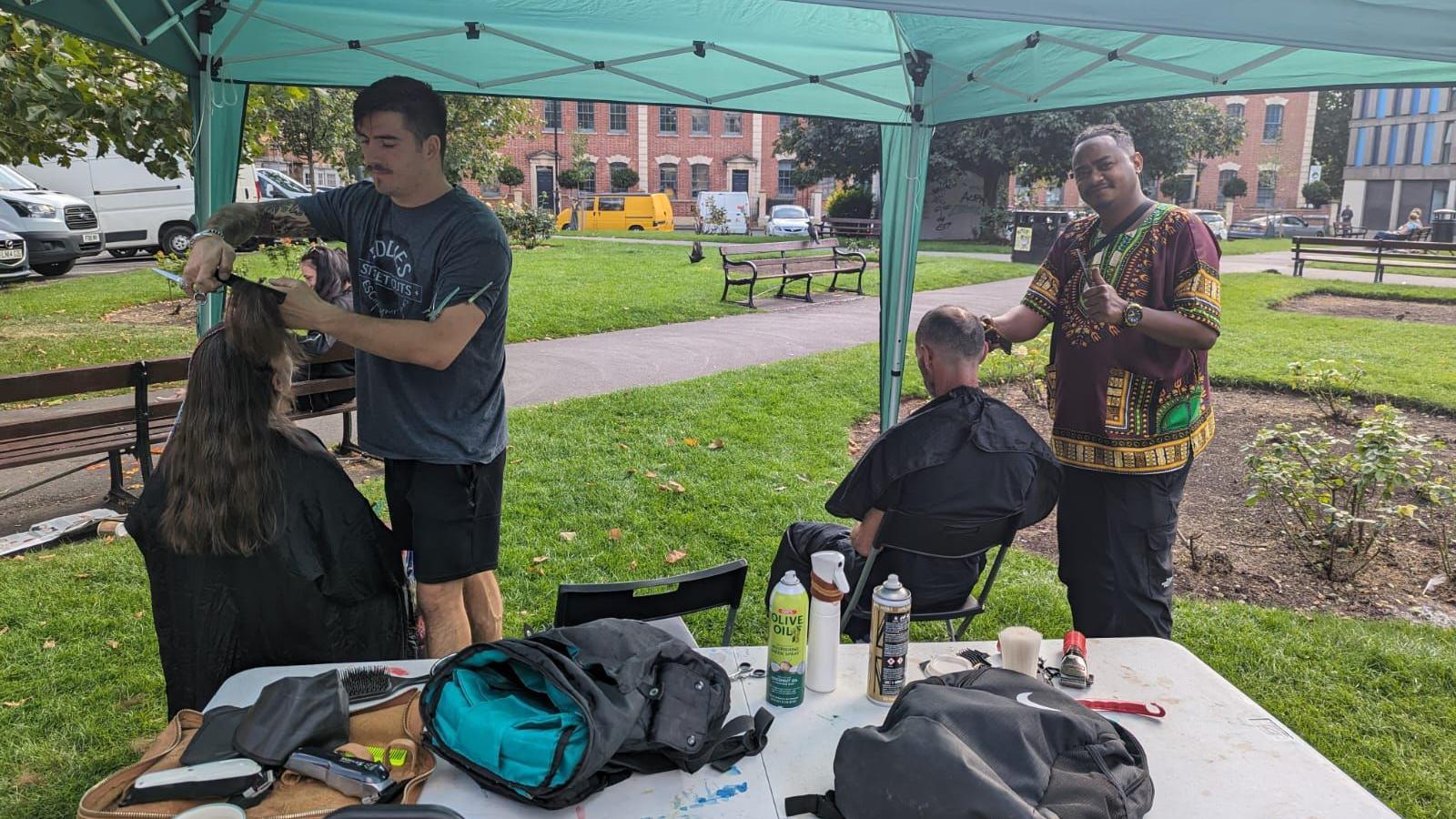 Two men cutting hair outside under a marquee