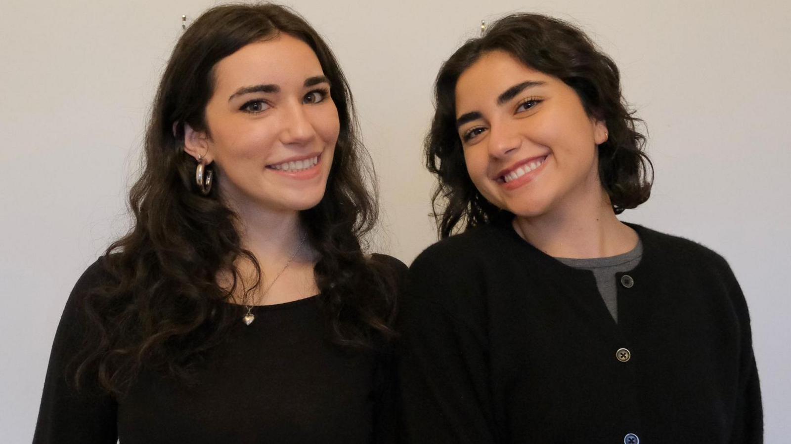 Two girls smiling at the camera against a white background. The girl on the left has long, dark, curly hair and is wearing a black long-sleeved top and silver jewellery. The girl on the right has shoulder-length, dark curly hair and is wearing a black cardigan and grey top. 