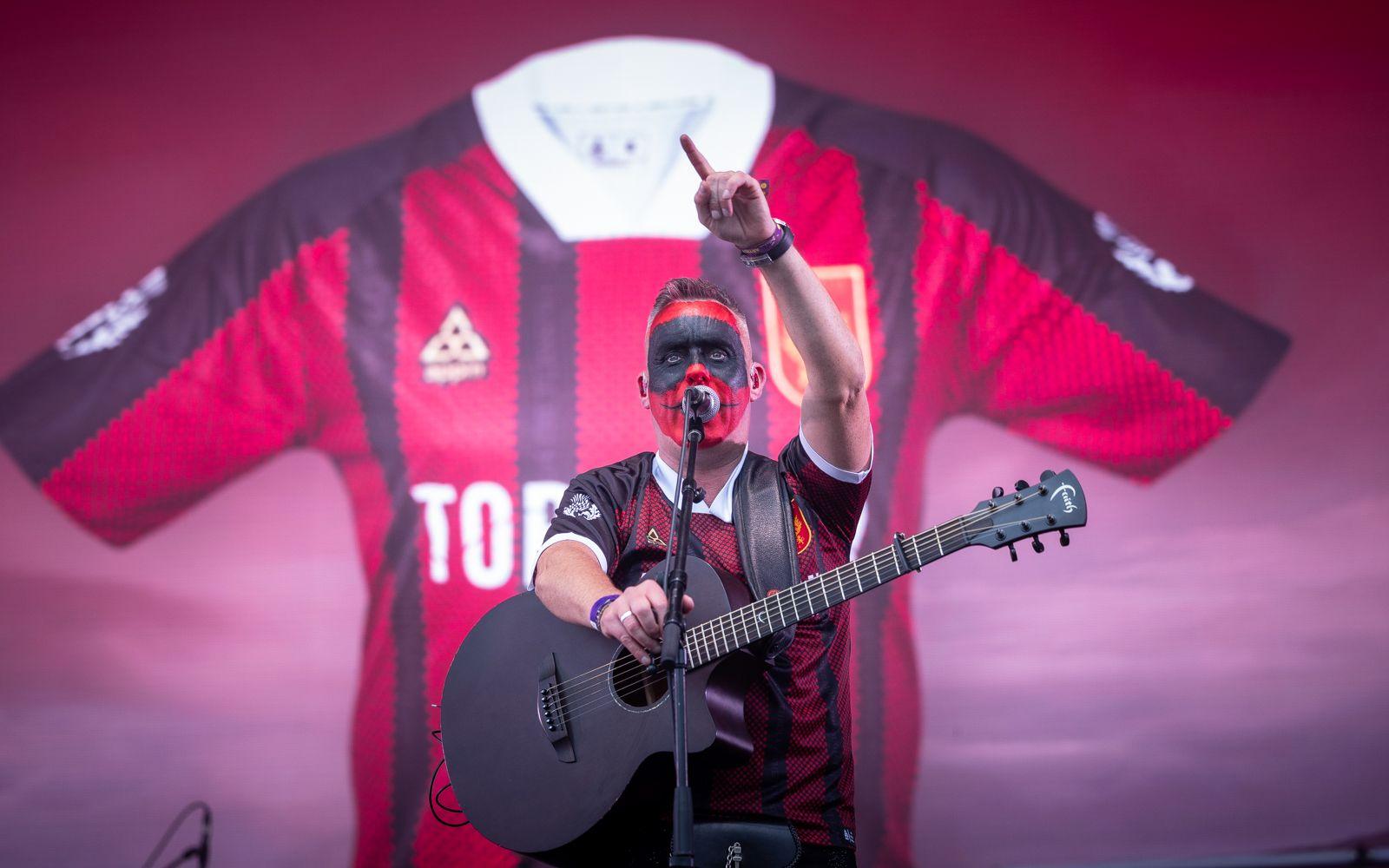 Torridon singer wearing red and black Inverness Thistle football top with his face painted red and black on stage, carrying a black guitar