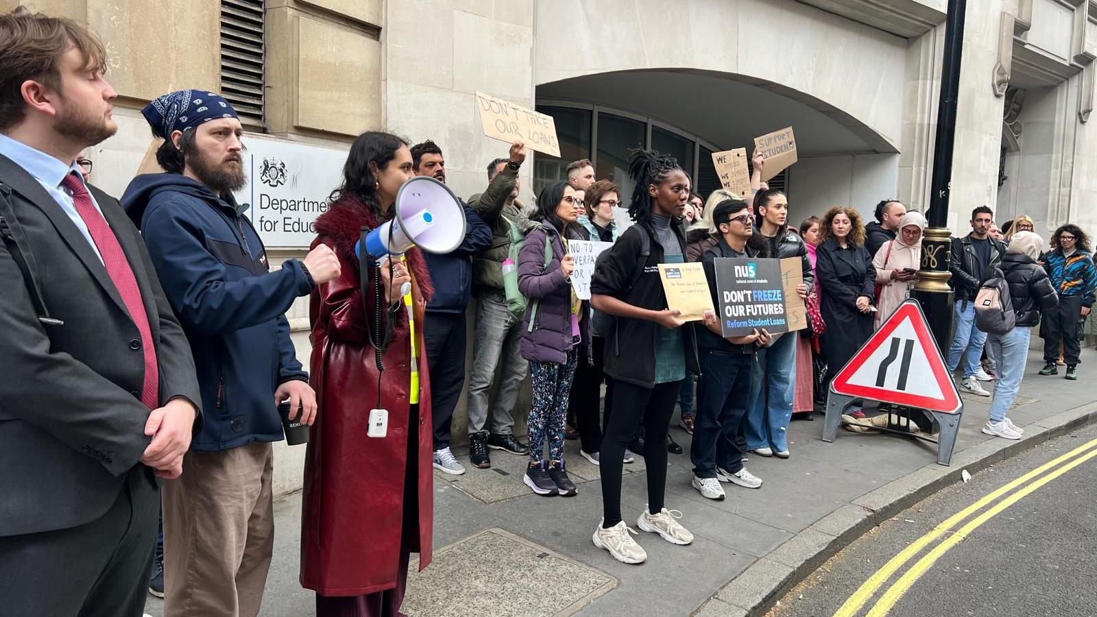 A line of protesters stand outside the Department for Education building, some holding placards and one using a megaphone. Visible signs include “Don’t hike our future student loans”. The group lines the pavement beside a road with temporary traffic signs.