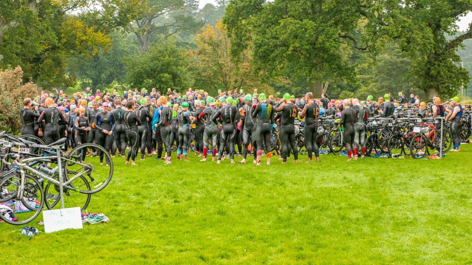 Dozens of people in wetsuits stand in a field. There are a number of bicycles. In the distance, there are a number of trees. 