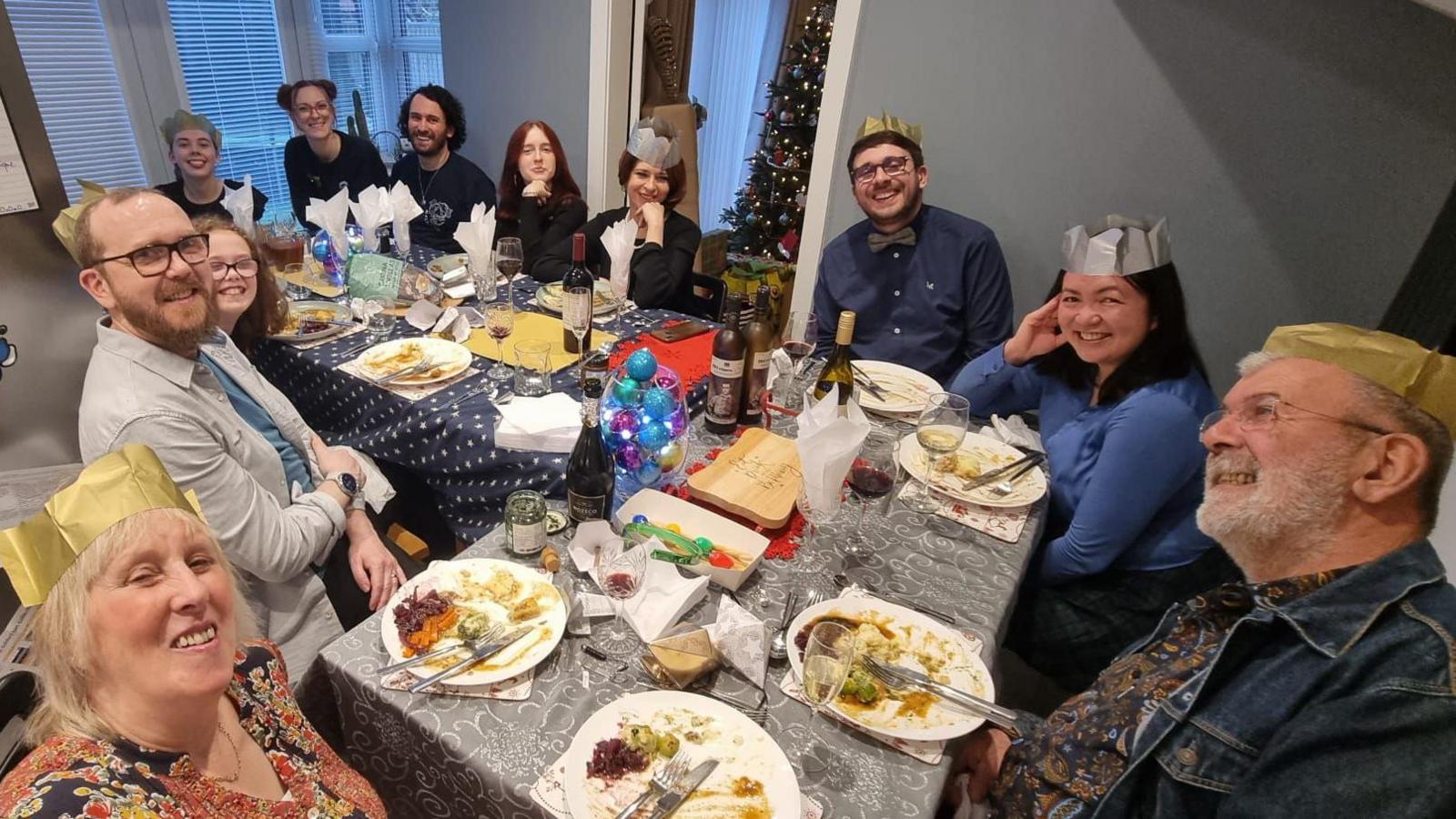 Eleven people sitting around an L-shaped table, which is covered with a cloth and has plates of Christmas dinner placed in front of all the diners. Seven are wearing paper gold-coloured crowns. There is a decorated Christmas tree in the corner. All are smiling at the camera.