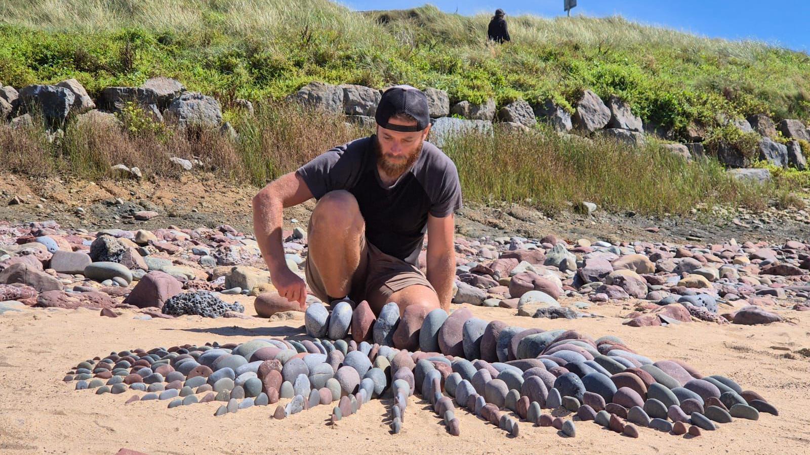 Man crouched down on the sand of the beach with a collection of stones carefully placed in size order.