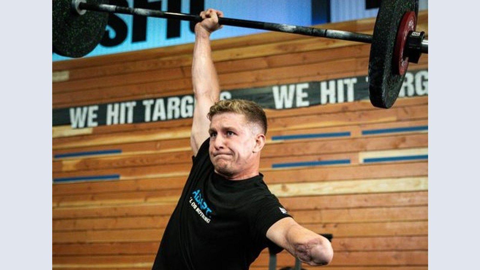 Henry Dunn in the gym lifting a weight above his head. His amputated arm is visible and he is wearing a black T-shirt and clearly exerting himself to lift the weight