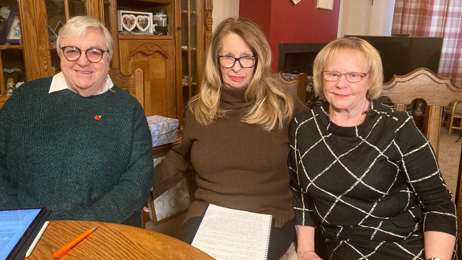 Three women sat at a living room table. One is wearing a green jumper and has short white hair with glasses, the one in the middle is wearing a brown turtle neck and has long blonde hair and also is wearing glasses. The other woman has short blonde hair and a black and white top. 