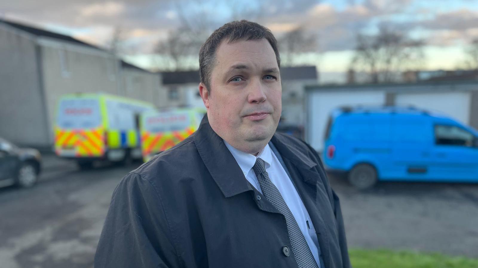 Councillor Craig Meek looking straight at the camera. He has dark hair and is wearing a dark jacket over a white shirt and brown tie. He is standing outside. In the background, blurred police vans and a blue van can be seen parked in front of houses and garages.