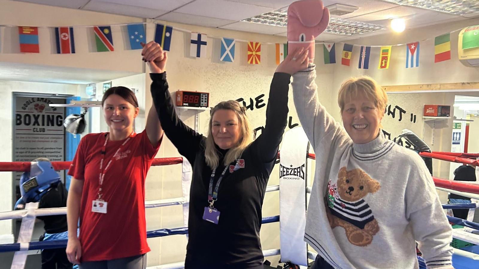 Three women in a boxing ring with their hands held aloft. India is wearing a red t-shirt and has brown hair in a ponytail. Debbie is in the middle with a long-sleeve black top on and has shoulder-length blonde hair, while Victoria has pink boxing gloves on and a white jumper with a teddy bear on it. She has short blonde hair. Outside of the ring, posters are on the wall and flags of different countries are hanging in a row along the top of the wall.