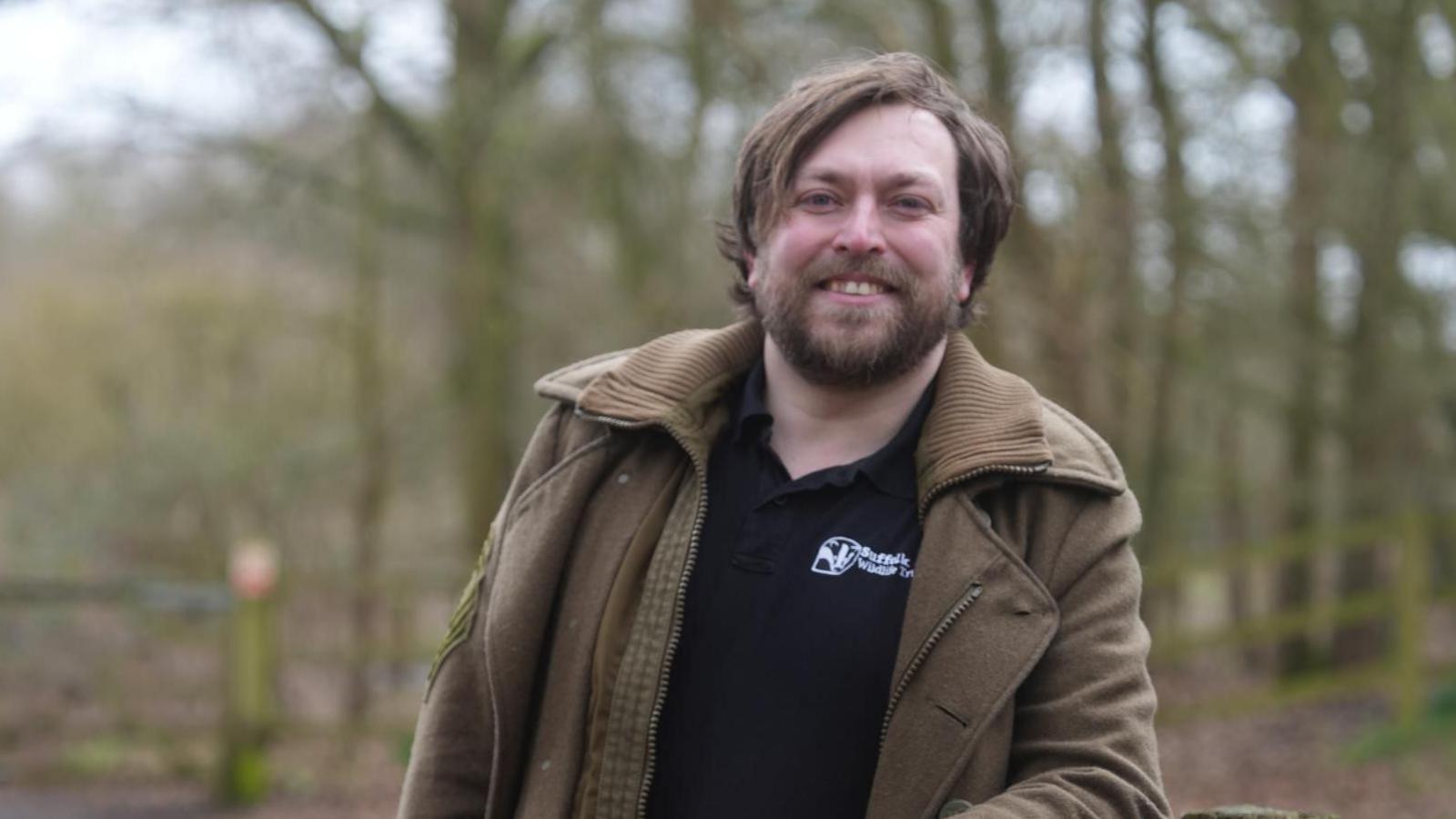 A man, Daniel Wade is standing in Fenland wearing a Suffolk Wildlife Trust t-shirt and army green coloured jacket. He has a beard and is smiling at the camera. 