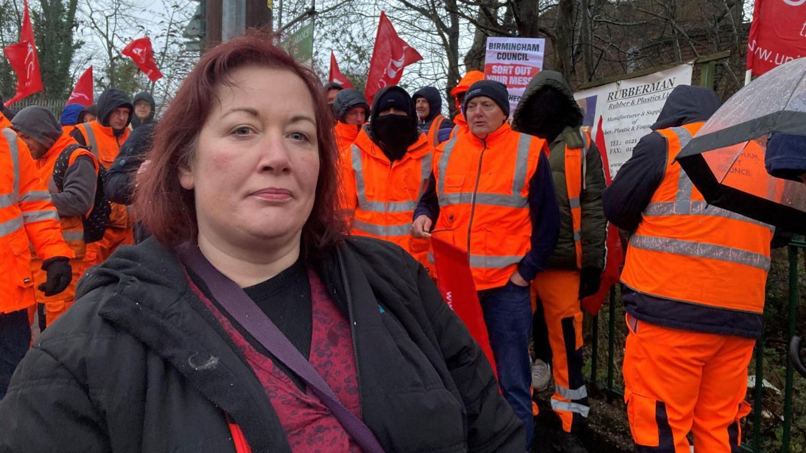 A woman stands in front of a group of men who are wearing high-vis orange jackets and waving red flags. The woman has short dark red hair, and she is wearing a black jacket over a red and purple top.