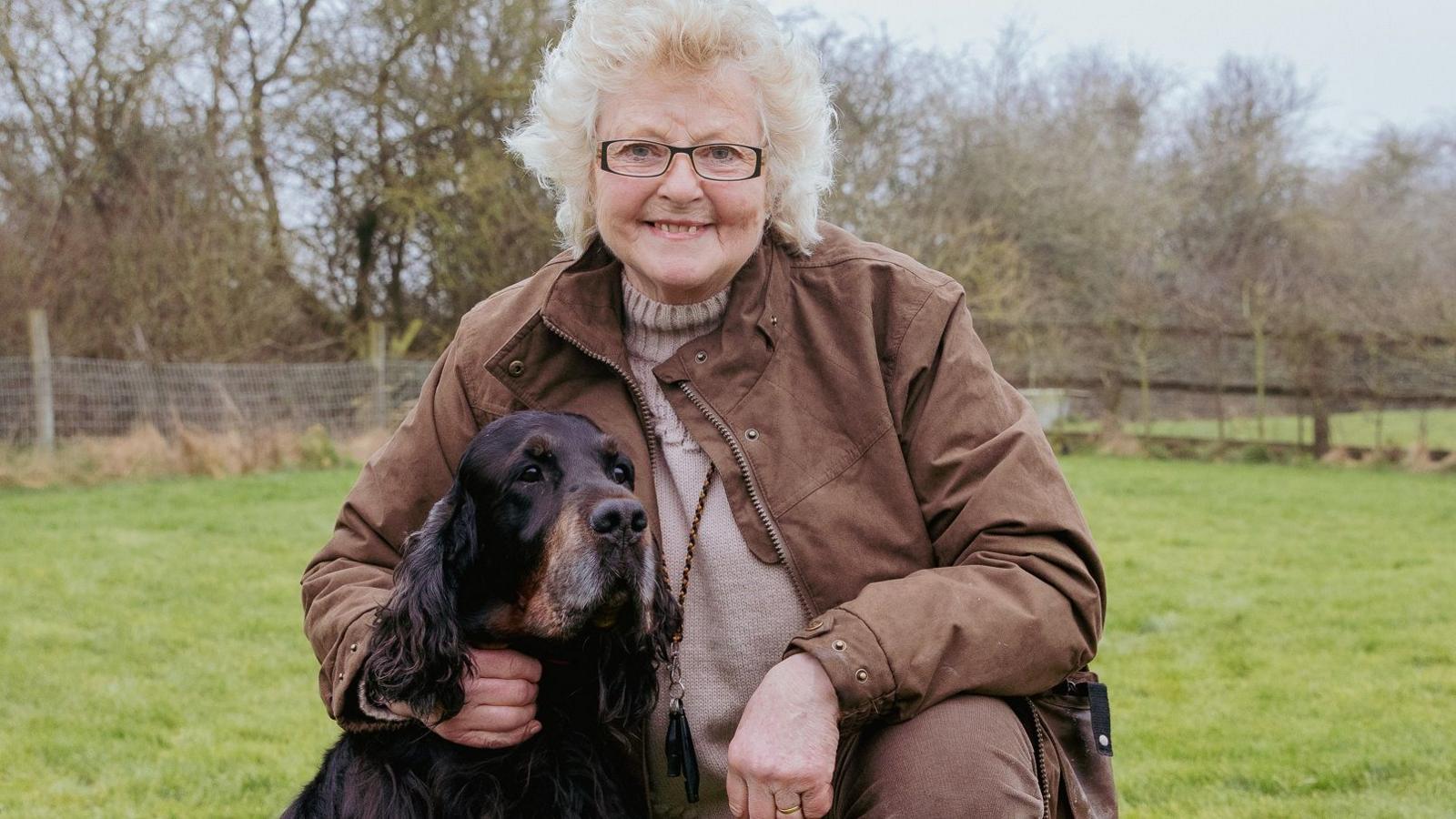 A woman with white curly hair is wearing a brown jacket with her arm around a Gordon Setter. They are both sat in a green field.