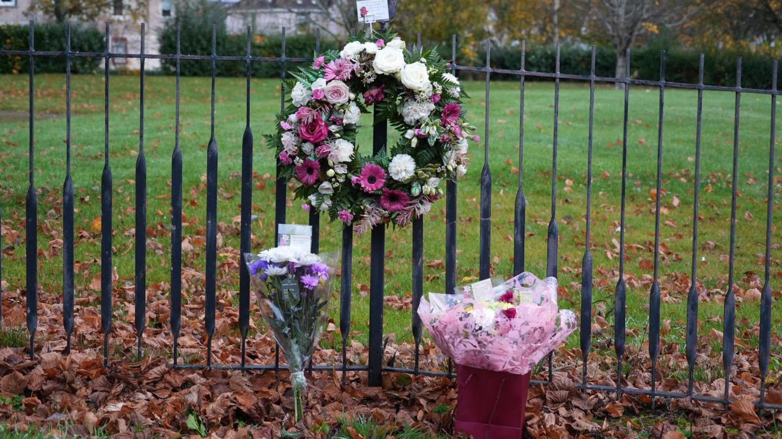 A wreath and two bunches of flowers left against a fence in Bellshill. Behind the fence is a stretch of grass, a hedgerow and a number of trees