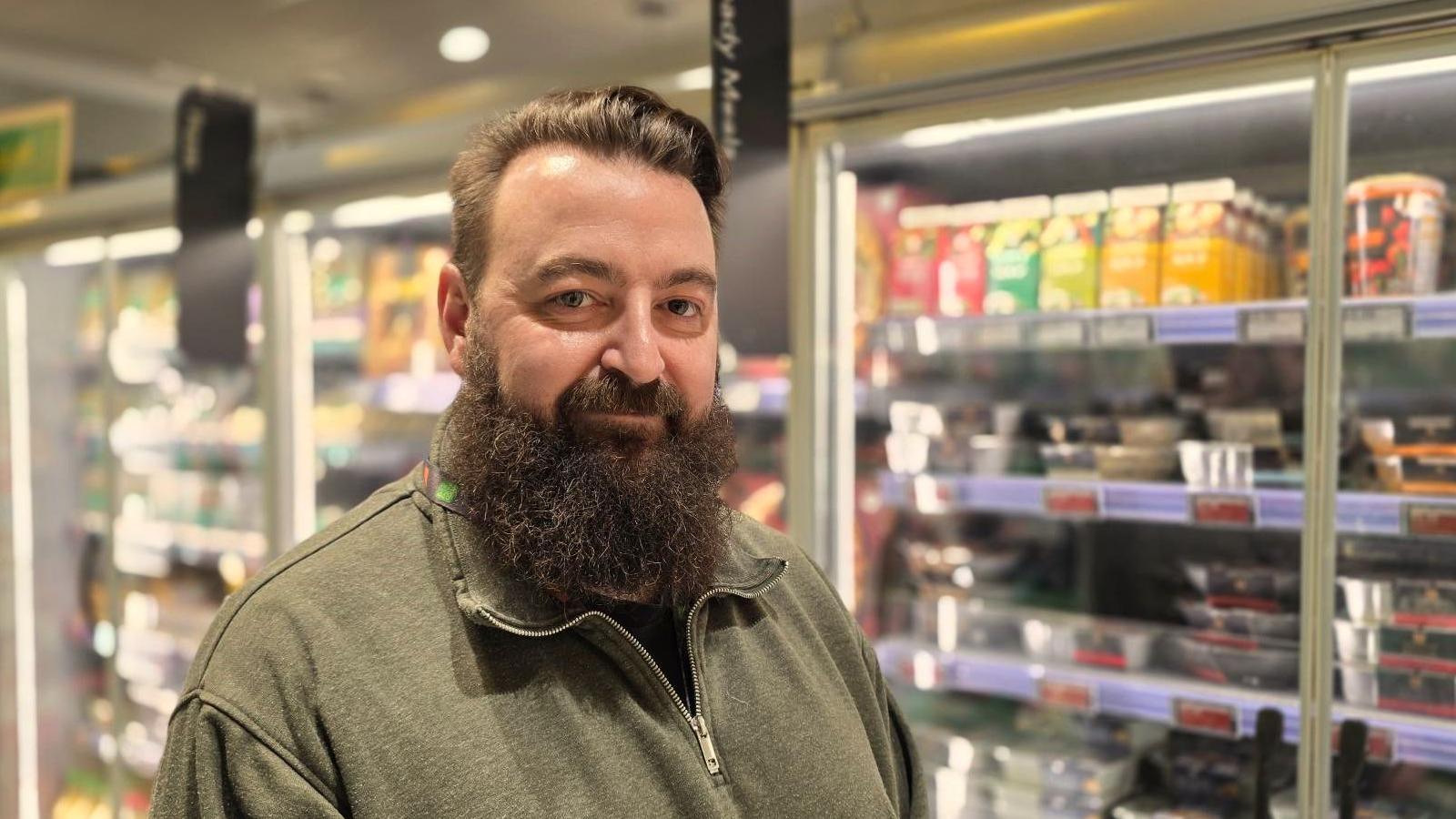 Lee Hammond in a supermarket aisle with stocked fridges behind him. He has a dark, bushy bear and is wearing an olive green zip-up top.