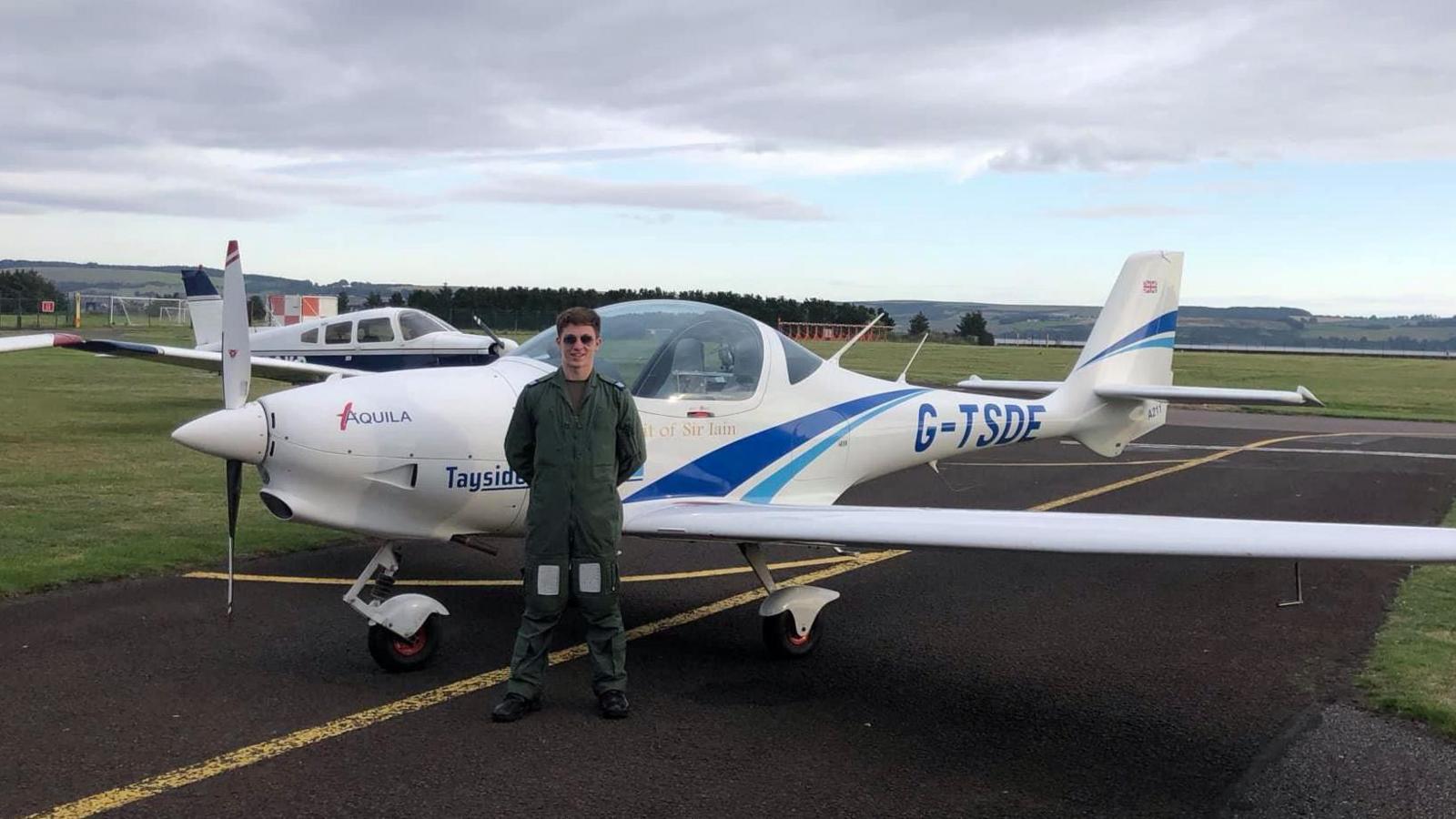 The 16 year old pilot, wearing a green flying suit, stands in front of a white two seater plane which is parked just off the runway of a small airfield.