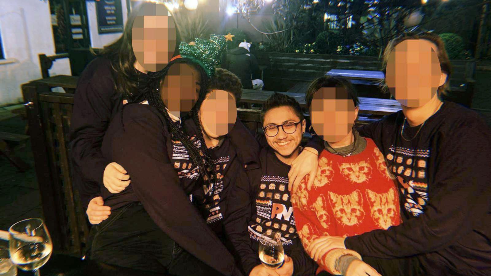 Stephan Kyriacou, who has dark hair, a beard and glasses, sitting with a group of other people who's faces we have pixelated. Several of them are wearing jumpers with the PinkNews logo, pixel art-style faces and a motif of a unicorn leaving a rainbow in its wake. Another person is wearing a red jumper with woven images of cats in yellow wool. A few white wine glasses are in shot.