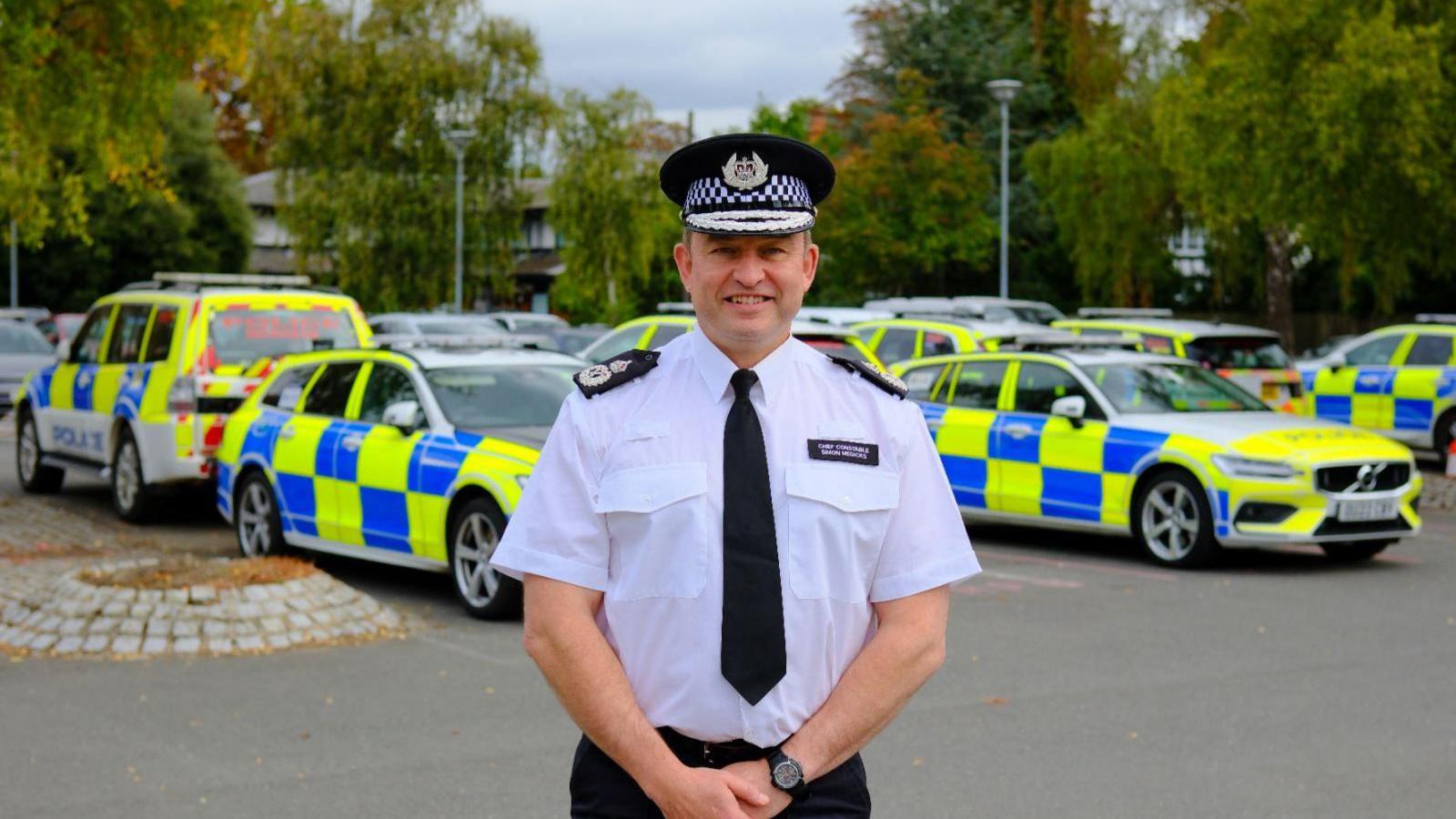 Simon wearing white police shirt and black trousers and a black tie and Black police hat- standing in front of police cars.