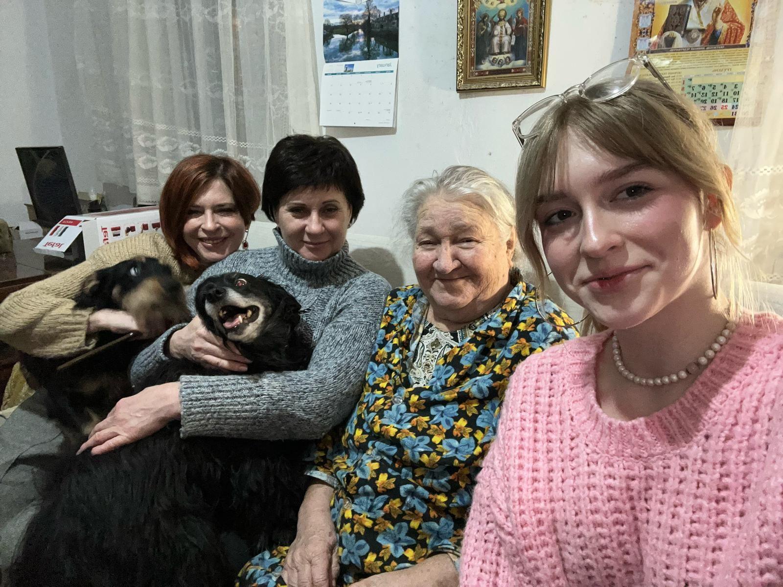 Four women sitting on a sofa. The one on the far right has blonde hair and is wearing a pink knitted top, a pearl necklace and plastic-framed specs on top of her head. The woman next to her on the left is much older and has grey hair, which is tied back. She is wearing a black, blue and yellow floral dress. The woman on the far left has chestnut-brown coloured bobbed hair and is wearing a beige jumper while holding a black dog on her lap. The woman sitting next to her on the right has black short-cropped hair and is wearing a grey turtle-neck jumper while stroking a black dog, which is on her lap.