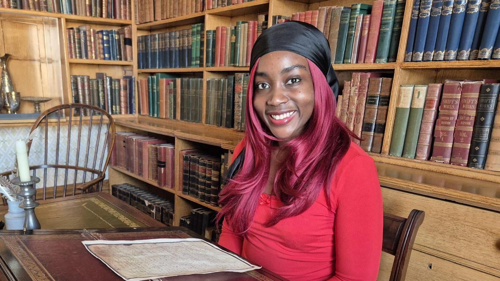 Princess, wearing a black head bandana and red top, sits at a antique table in the library with shelves of books behind her.