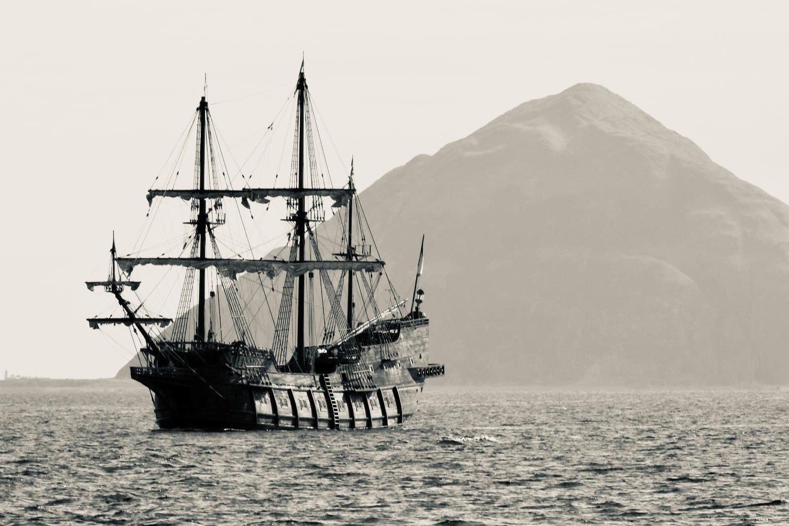 A replica galleon with tall masts at sea, with a large island in the background.