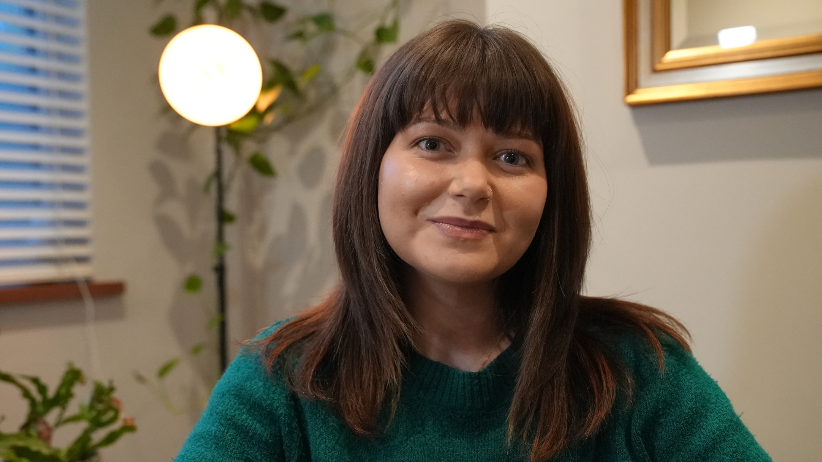 Një grua e veshur me një pulovër të thurur ngjyrë jeshile të errët është ulur brenda. Ajo ka flokë të drejtë ngjyrë kafe me gjatësi mesatare dhe skaje më të çelëta. Në sfond, një llambë e rrumbullakët e ngrohtë ndriçon pranë një bime me gjethe. Një dritare me perde horizontale është e dukshme në të majtë dhe një pasqyrë me kornizë varet në mur në të djathtë.