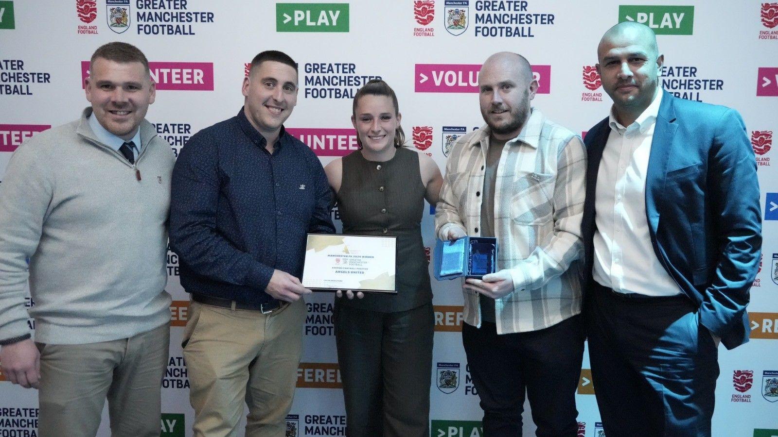 Olly Monk, wearing a checked shirt, is beside footballer Ella Toone and members of the Football Association to recieve a framed award.