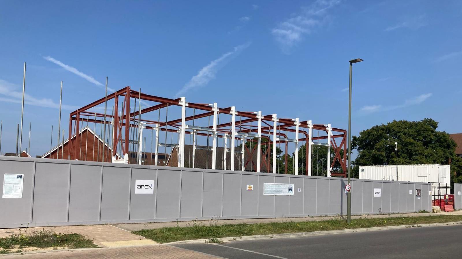 Grey hoarding surrounds development which is steel beams and bars surrounded by scaffolding