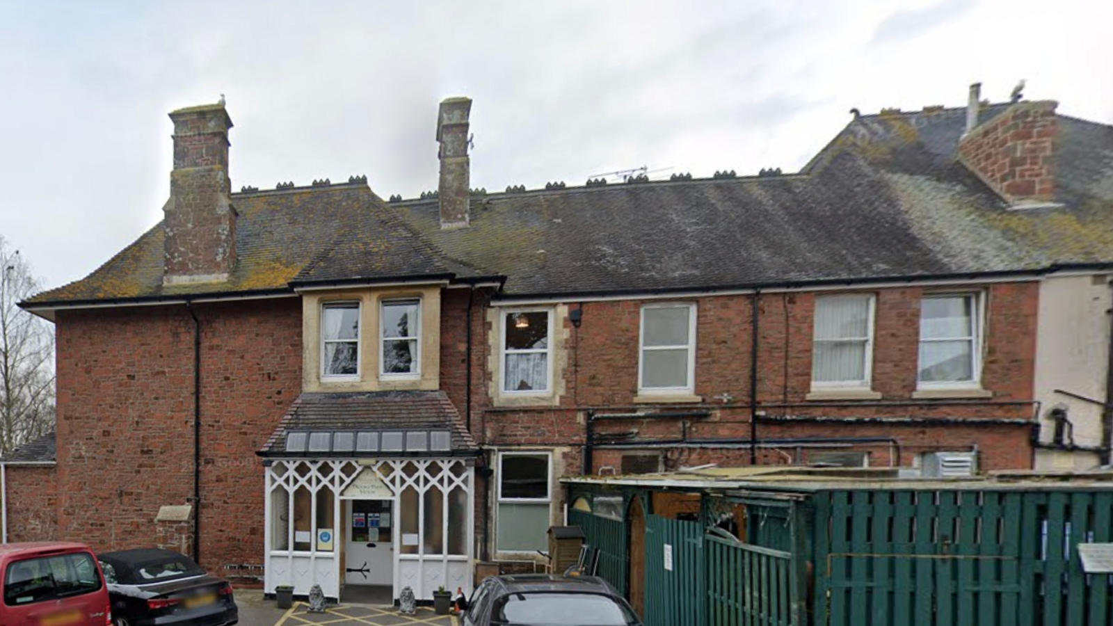 A brick two storey building with a grey slated roof with three chimneys and a white porch, two red cars and two black cars parked outside on a grey sky day.