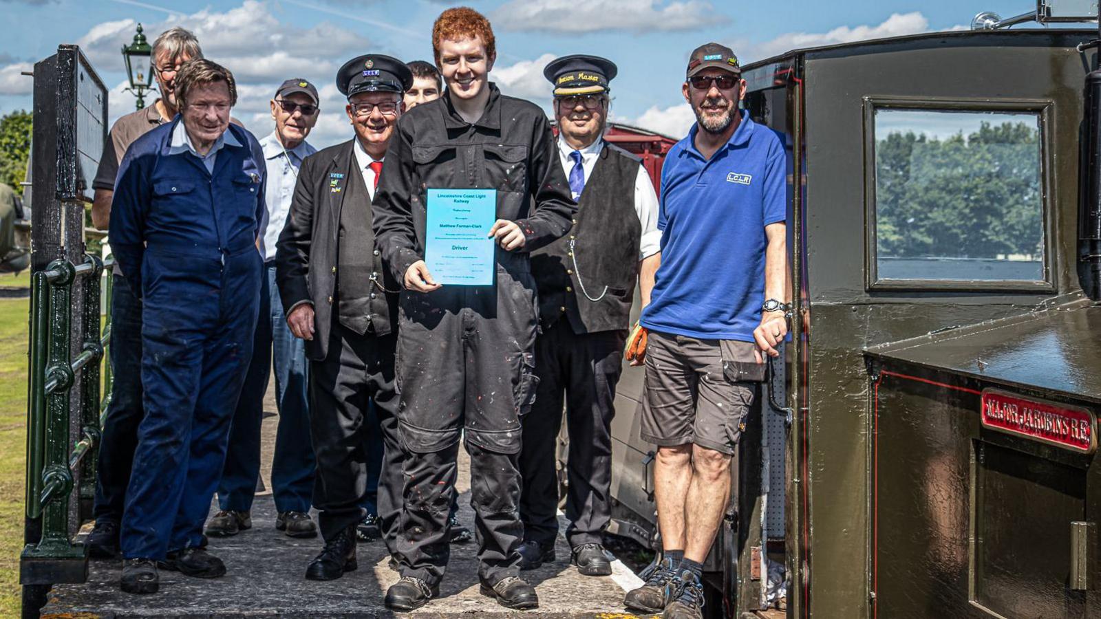 Mr Forman-Clark, holding his driver's certificate, stands on a platform next to seven other railway staff, some wearing uniforms comprising peaked caps