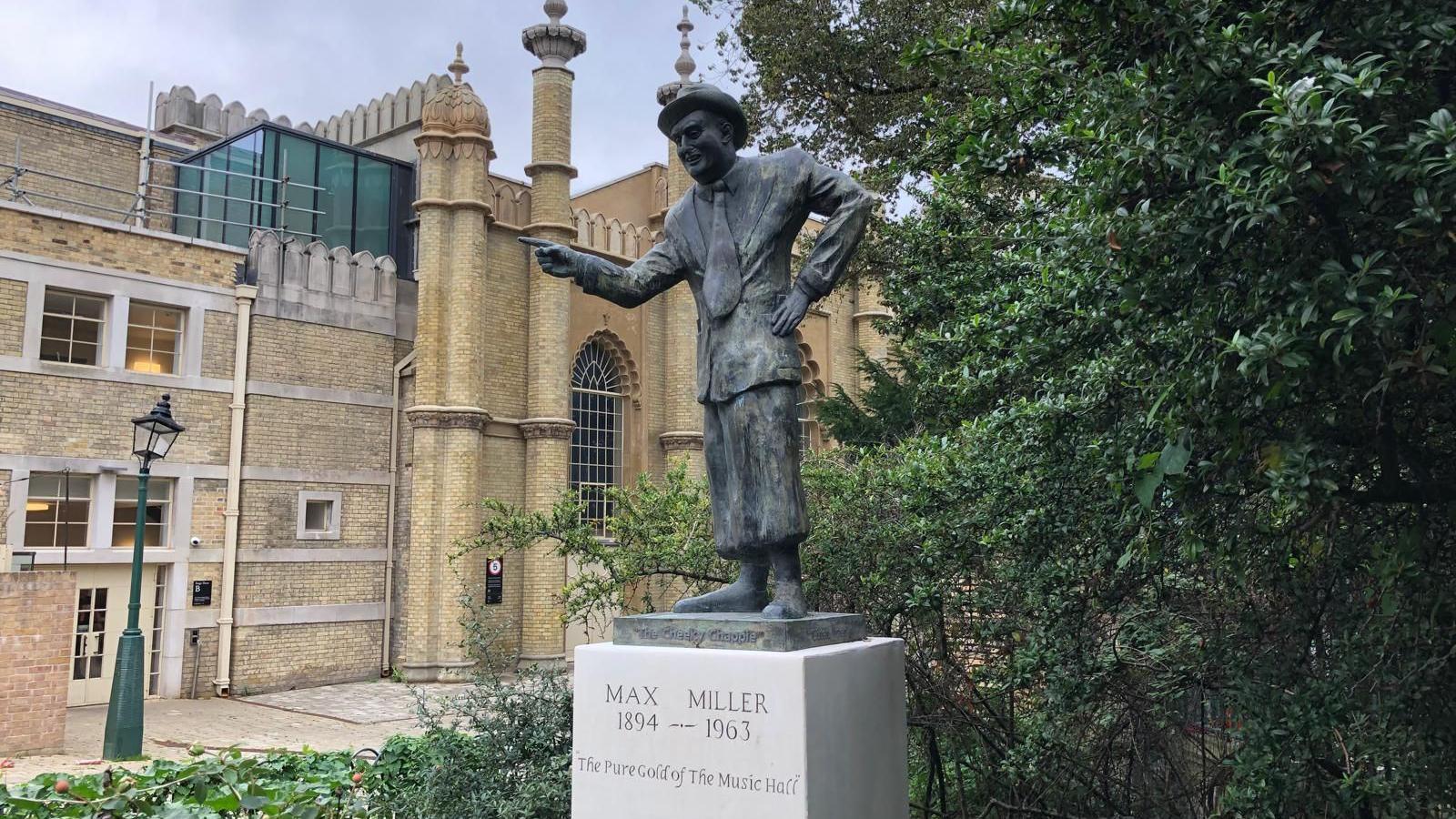 The statue of Max Miller in the Royal Pavilion Gardens, Brighton, with the Brighton Dome and a slightly bent lamppost in the background.