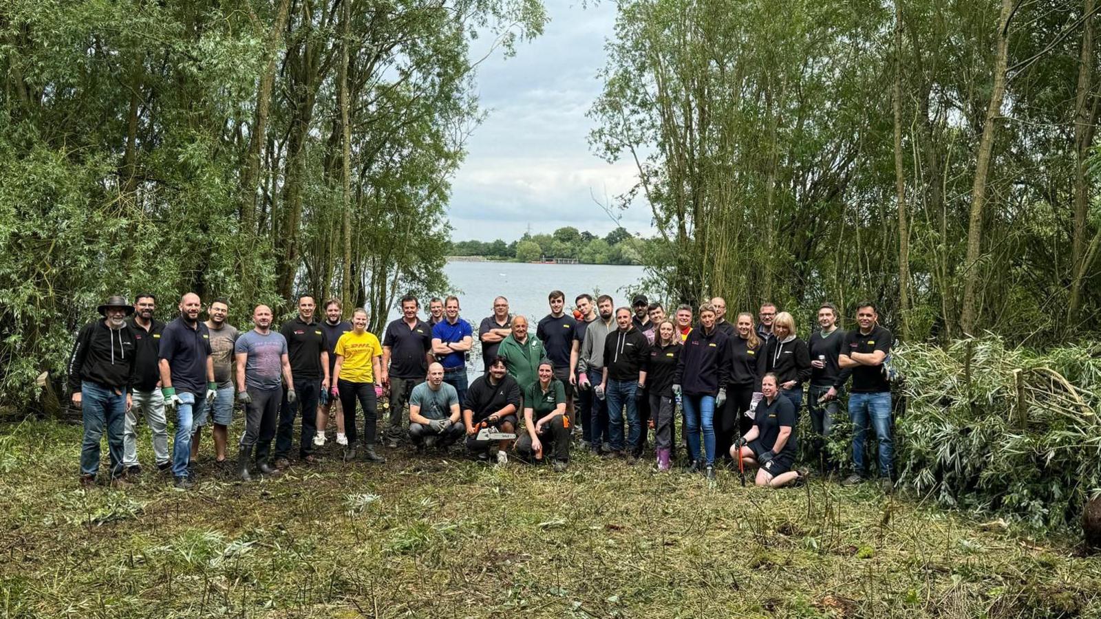 About thirty people standing or squatting in a semi-circle- there is a mixture of ages and genders. There is a grass and dirt surface in front of them, and a reservoir behind flanked by tall trees.