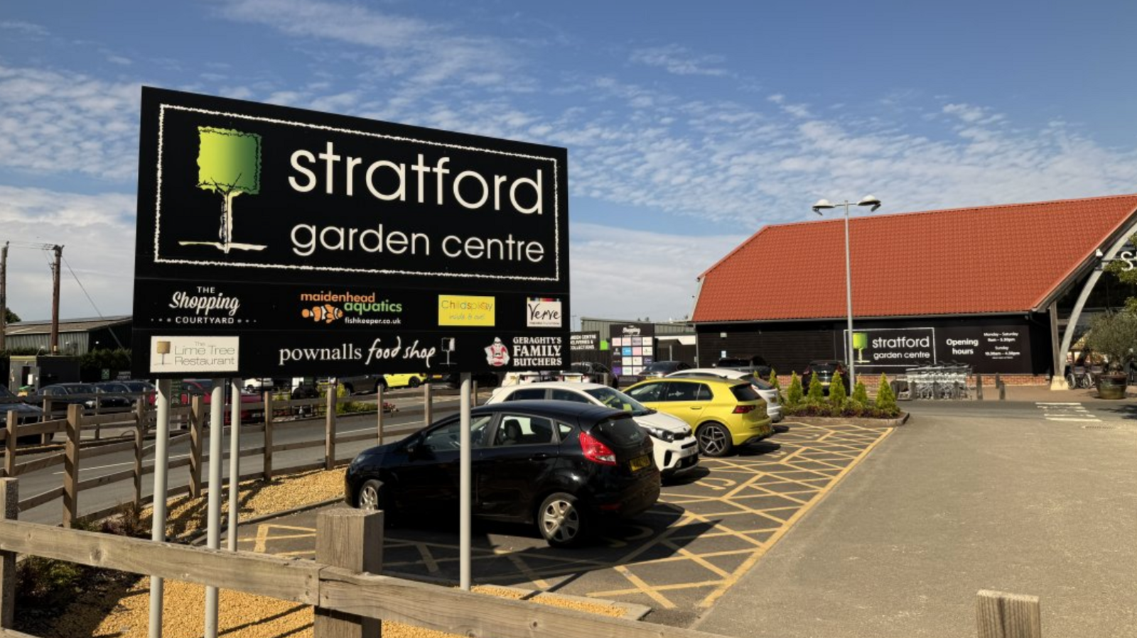 A sign for Stratford Garden Centre sits at the foreground of a carpark. The garden centre is at the far end of the carpark and has a large red roof and black facade. There are some cars parked up in disabled bays in front. It is sunny day.