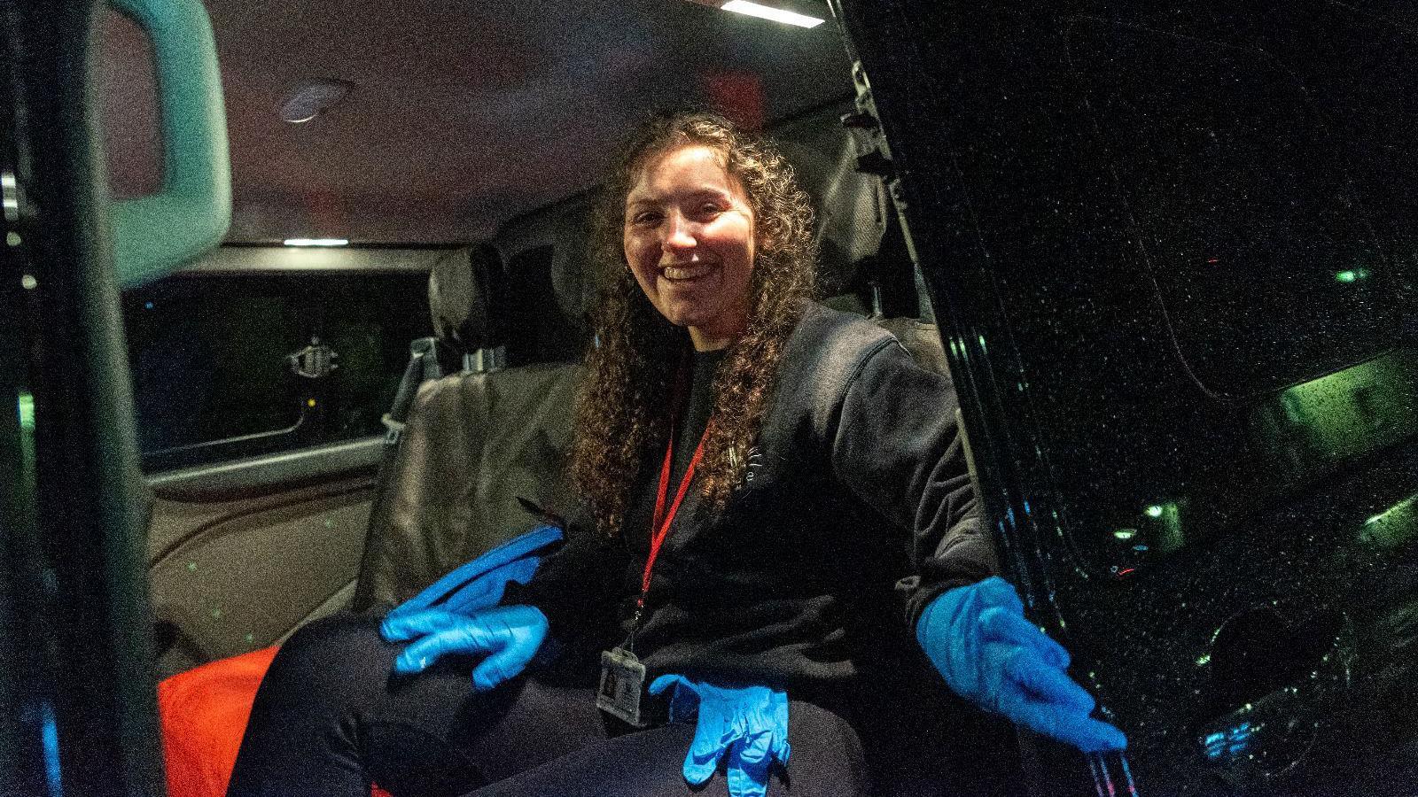 A woman sitting inside a vehicle wearing all black, with blue medical gloves on her hands and a spare pair on her lap. She has long curly brown hair and a red lanyard around her neck