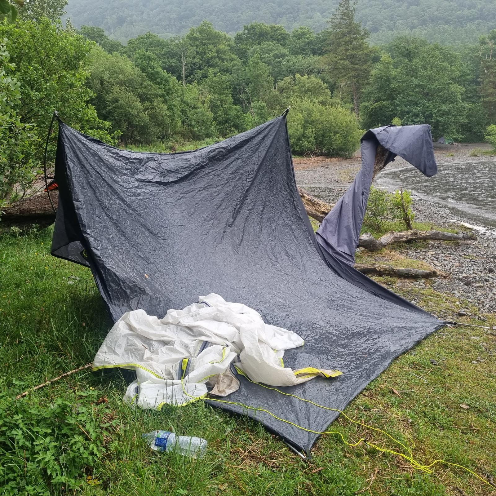 The remains of a tent abandoned in a wooded area. There are other items of litter including a white plastic sheet and an empty water bottle.