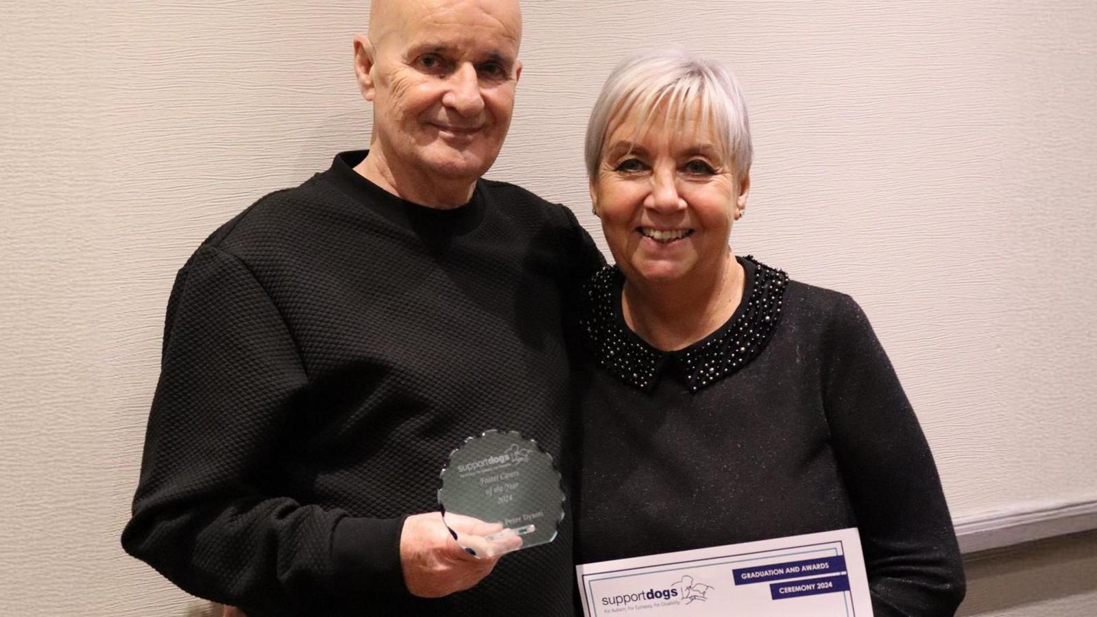 Janet and Peter are stood together with arms around each other. They're holding a certificate and a small, glass trophy.