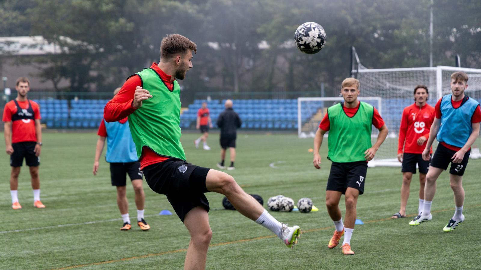 A man kicks a football in the air on a green pitch. Other players in green and blue bibs look on, with two goals in the distance.