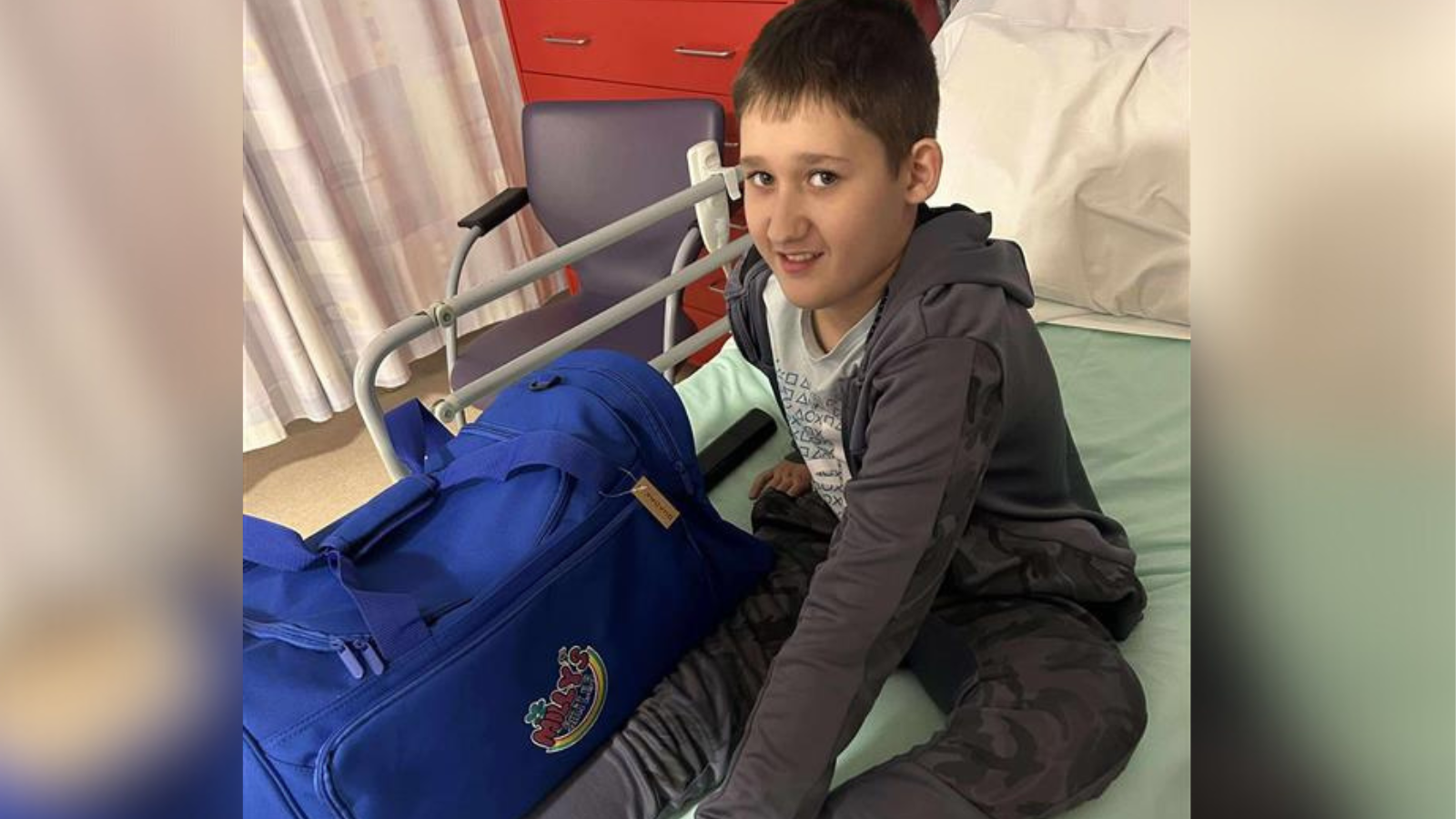 Archie sits on a hospital bed, next to a large overnight bag. He wears grey tracksuit top and bottoms, and has short brown hair.