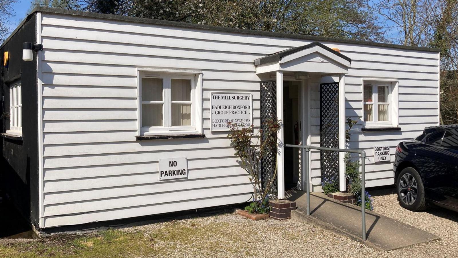 The single-storey surgery building pictured from further back. A gravel drive leads to its entrance, which has a black trellis and plants either side of the door. The building's sides are black and the front is made of white weatherboards. A small ramp allows for wheelchair access to the door.