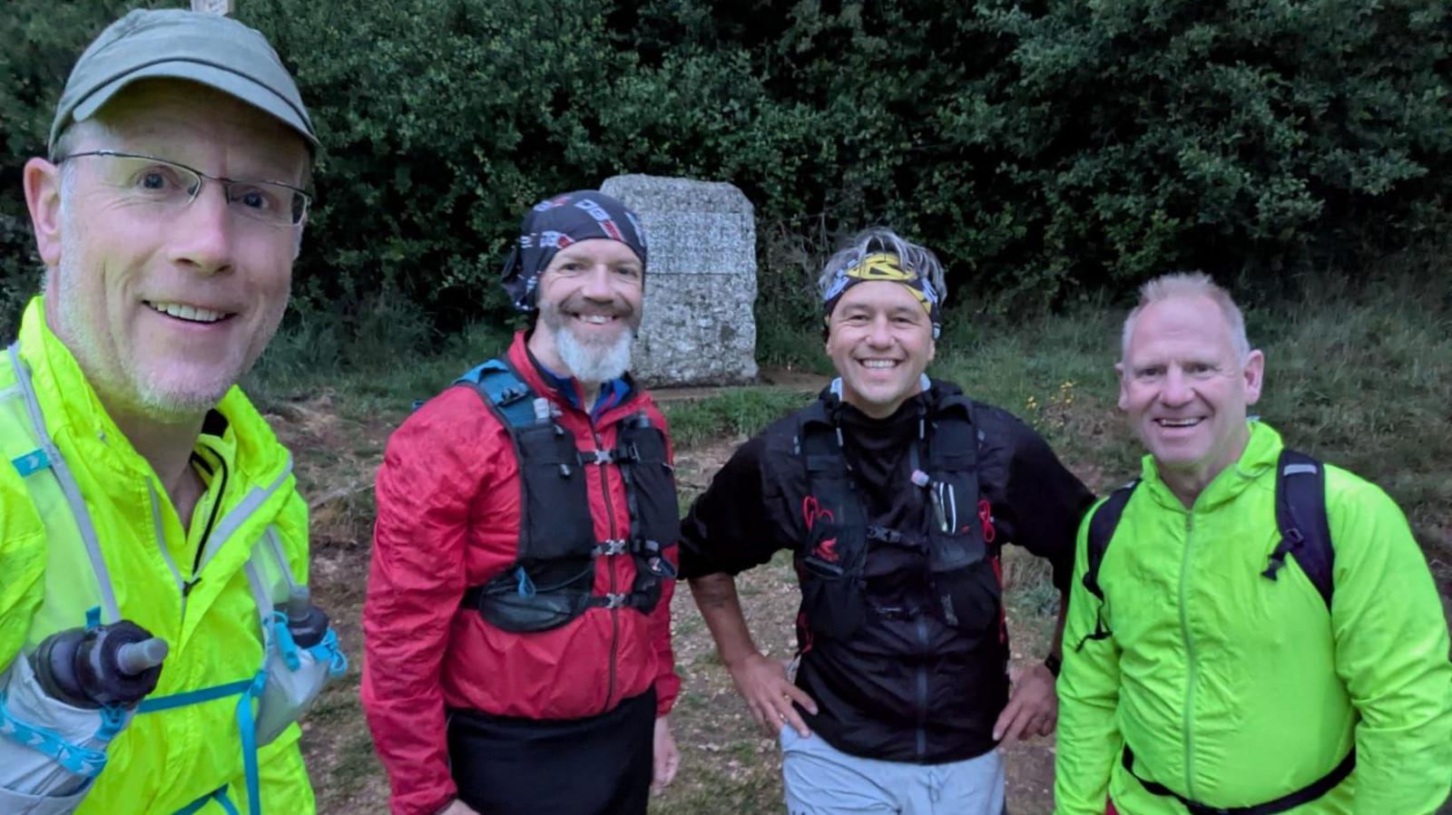 All four men smiling to the camera in a field. They are wearing bright waterproof clothing and have on running vests.