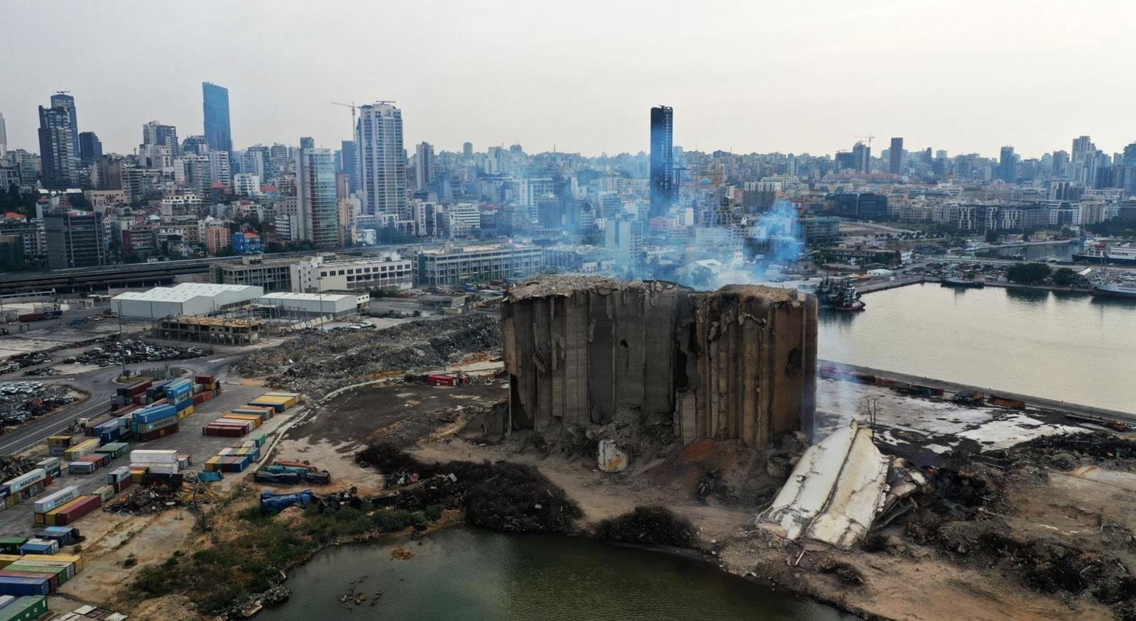 Remains of a storage building in Beirut which was destroyed in a large explosion. The severely damaged structure is surrounded by port facilities and shipping containers. The city of Beirut is in the background.