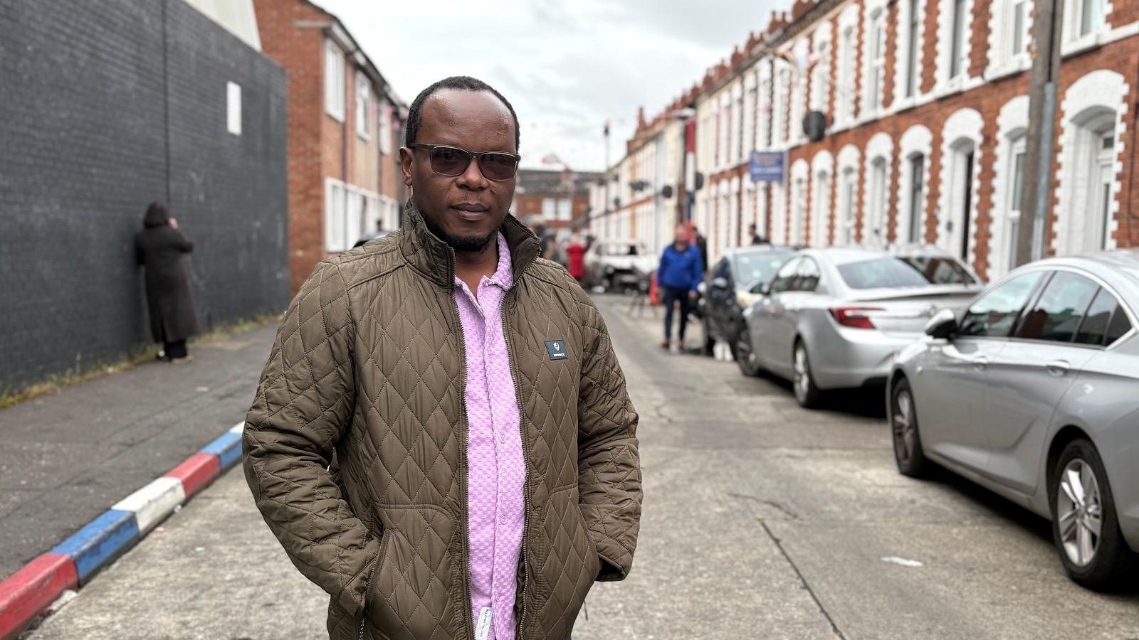 A man standing in a street lined with terraced houses. He is wearing a brown anorak and pink shirt and wearing dark-lensed glasses. He is balding, and has some hair towards the back of his head. On the right side there is a line of cars parked in front of the houses. On the left the kerbstones are painted red, white and blue. It is an overcast day. 