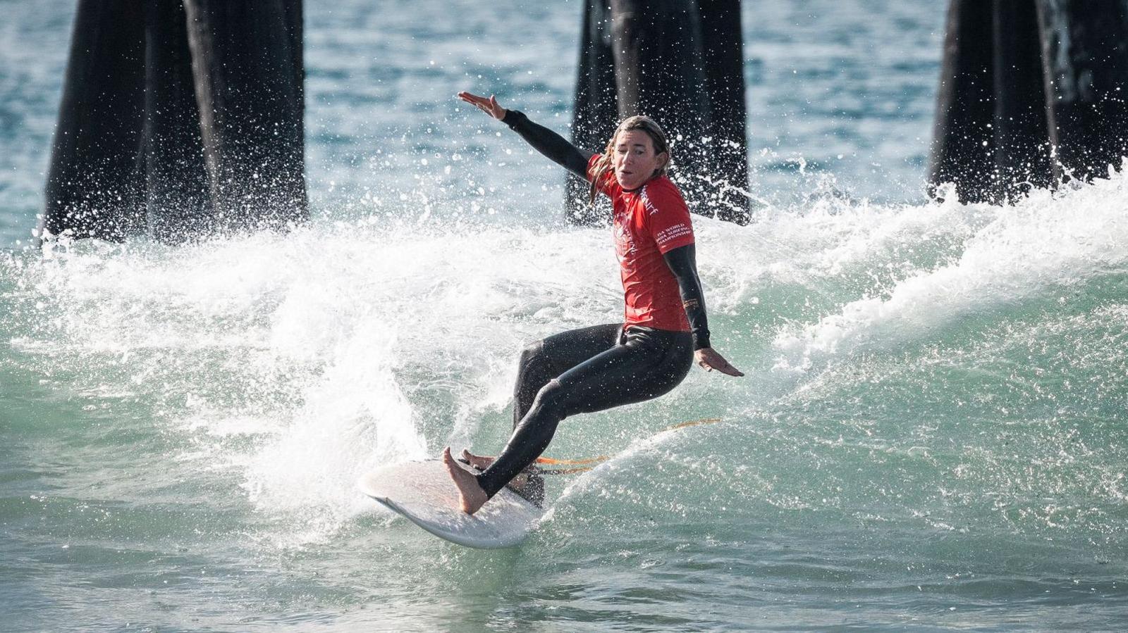 Melissa Reid is surfing with her arms outstretched and left foot balancing on her heel. She is wearing a Team England T-shirt over a wetsuit. Behind her is the pier and the water is clear.