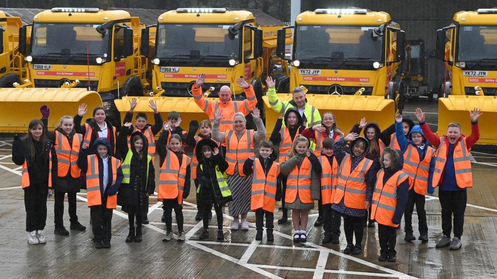 A group of school children wearing hi-vis jackets stand are waving at the camera. They are standing in front of a row of yellow gritters, which display their new names on red banners.