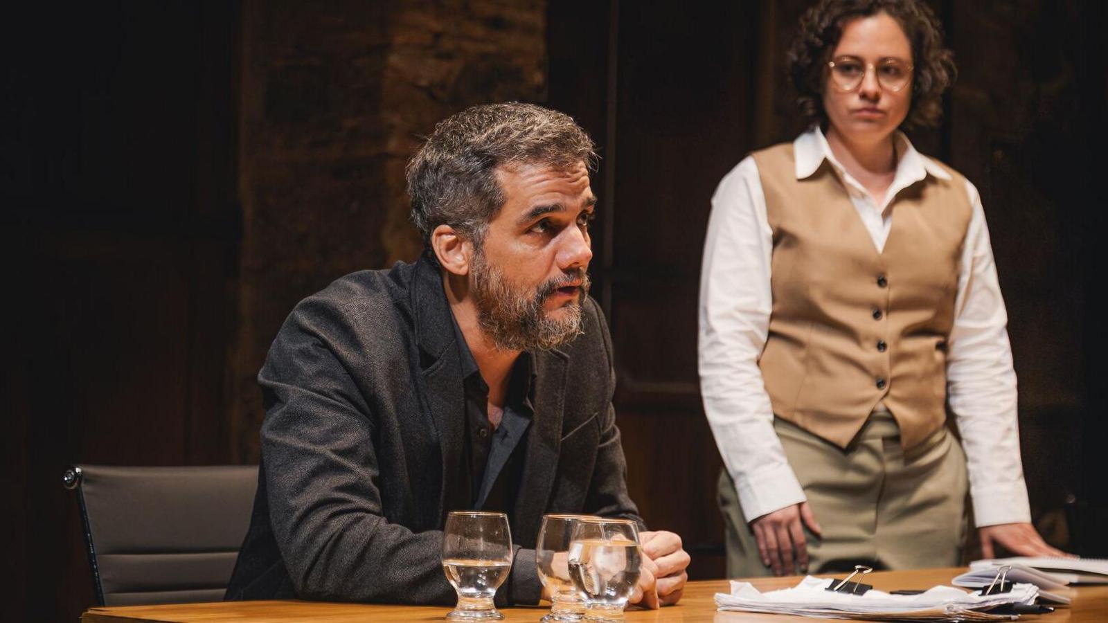 A man sitting at a table with a woman standing beside him. There are glasses containing water on the table and a stack of papers. The man has dark hair and a beard and is wearing a dark blazer over a dark shirt. He is leaning slightly forward with his arms resting on the table. The woman has short, curly hair and is wearing glasses. She is dressed in a white shirt and a tan waistcoat and trousers. 