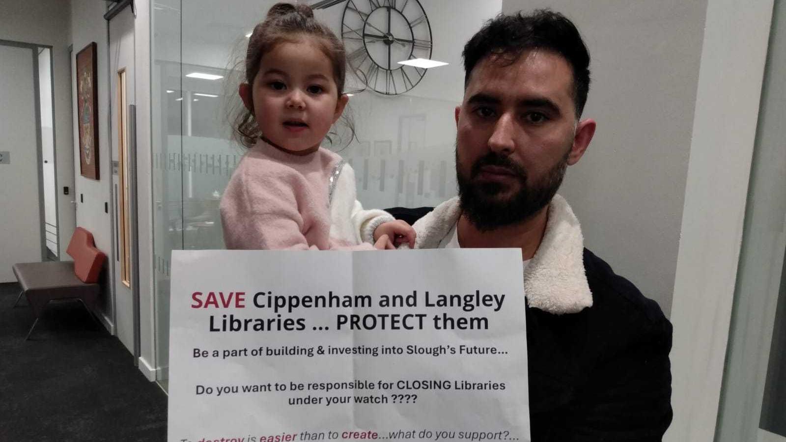 A man with dark hair and a beard, wearing a black coat and holding up a sign saying "Save Cippenham and Langley Libraries". He's holding a two-year-old girl with dark hair and wearing a pink jumper.