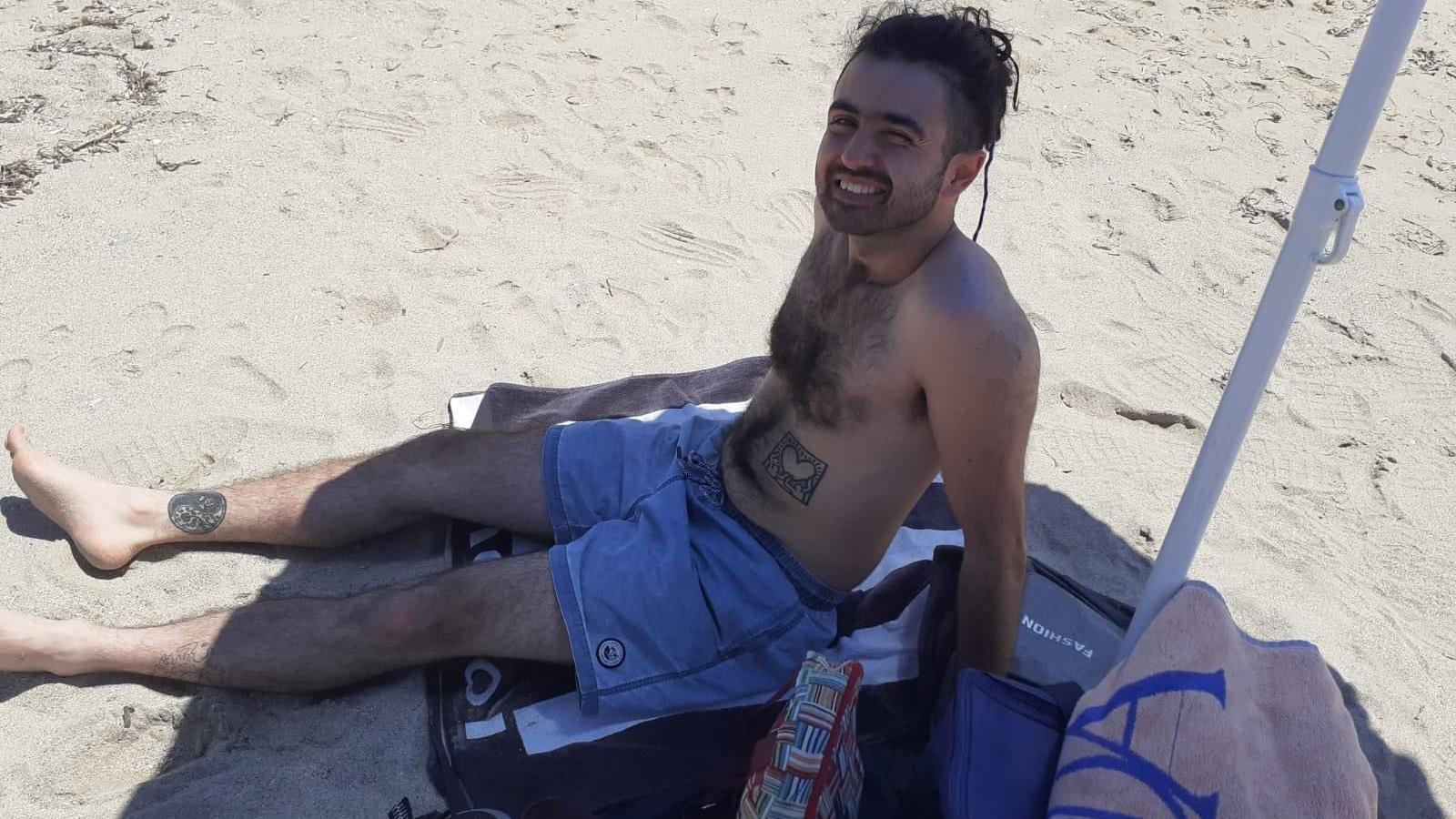 A man lying on a sandy beach wearing shorts, smiling up at the camera