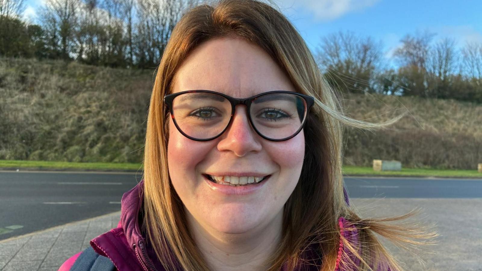 Caryl mewn sbectol a chôt binc yn gwenu at y camera