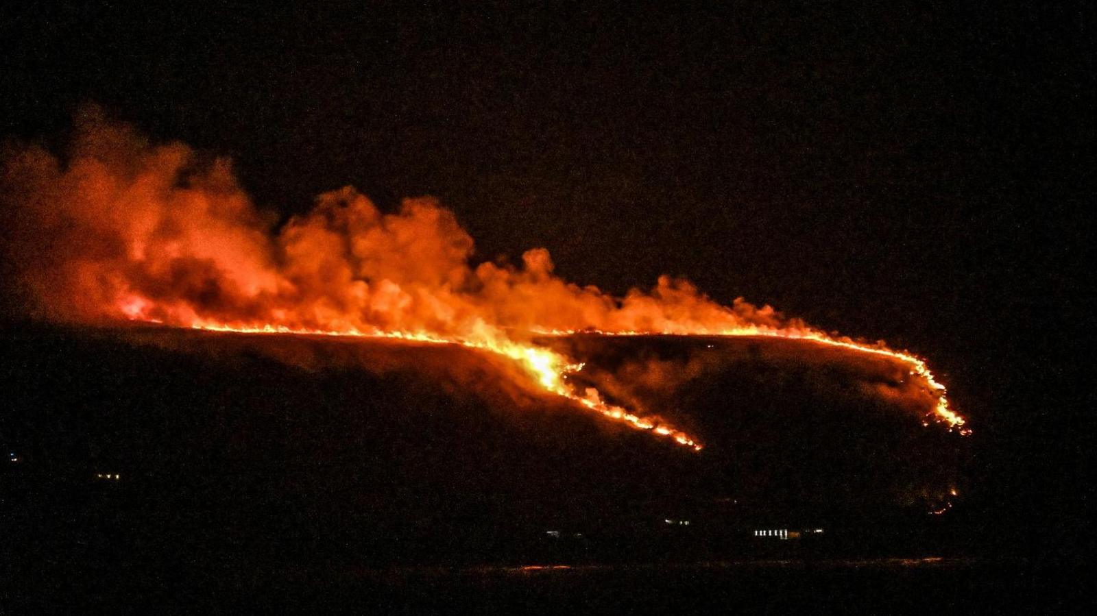 A dark view of a rural area with a long strip of large orange flames and billows of smoke on a hillside