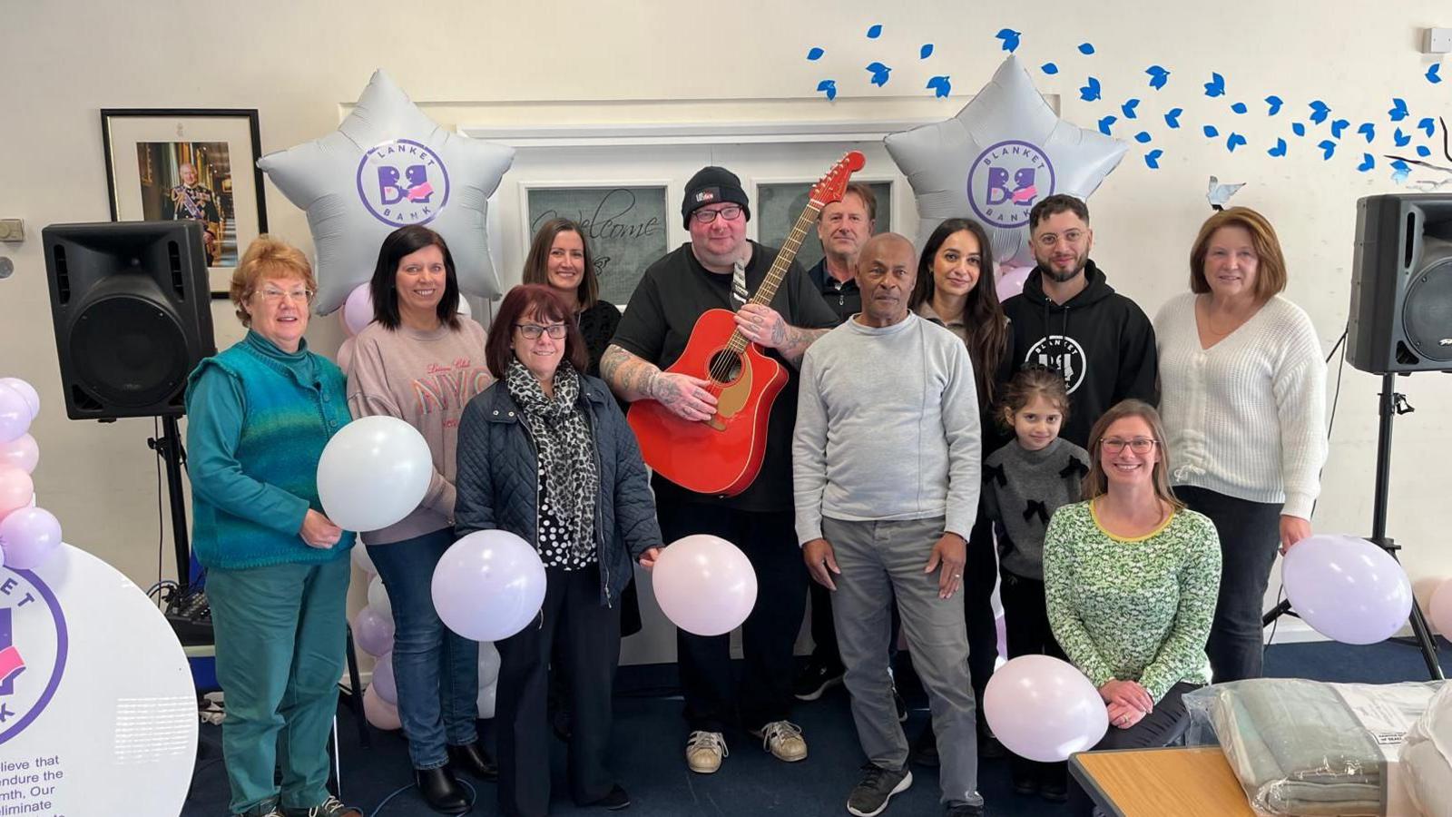 A group of people stand around a man with a guitar at the launch of the Blanket Bank. They hold balloons and are in front of speakers and double doors.